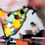 A student sits on an orange bench while using a laptop framed by colorful artwork.